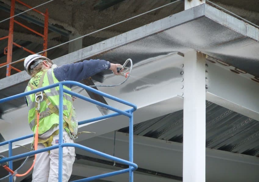 Aplicador em equipamento de proteção individual pulverizando tinta intumescente sobre viga e pilar metálicos em obra